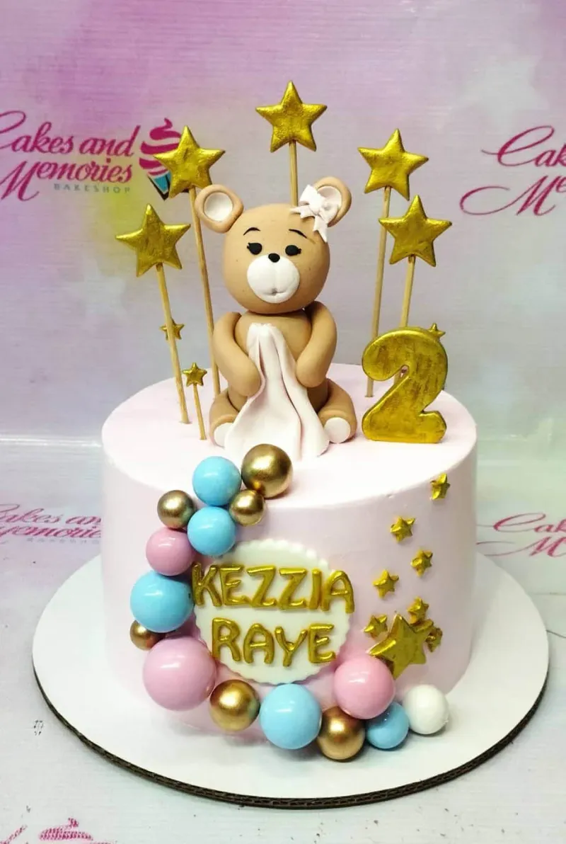 Pink 1-tier birthday cake featuring a 3D fondant bear with a white blanket, gold stars, and colorful spheres. Pink 1-tier birthday cake featuring a 3D fondant bear with a white blanket, gold stars, and colorful spheres.