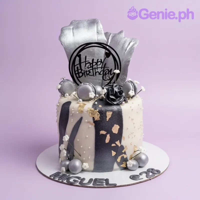A white, black, and silver 1-tier birthday cake featuring silver gumpaste sails, a black rose, silver macarons, and 'MIGUEL @ 28' lettering. A white, black, and silver 1-tier birthday cake featuring silver gumpaste sails, a black rose, silver macarons, and 'MIGUEL @ 28' lettering.