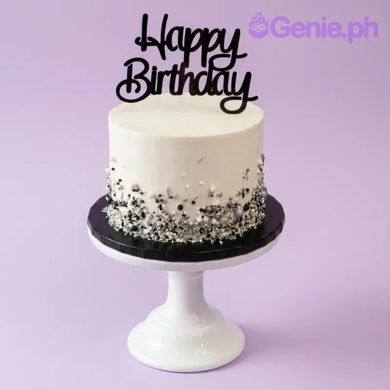 A white 1-tier cake on a black base board, featuring a black Happy Birthday script topper and silver, black, and white sprinkles. A white 1-tier cake on a black base board, featuring a black Happy Birthday script topper and silver, black, and white sprinkles.