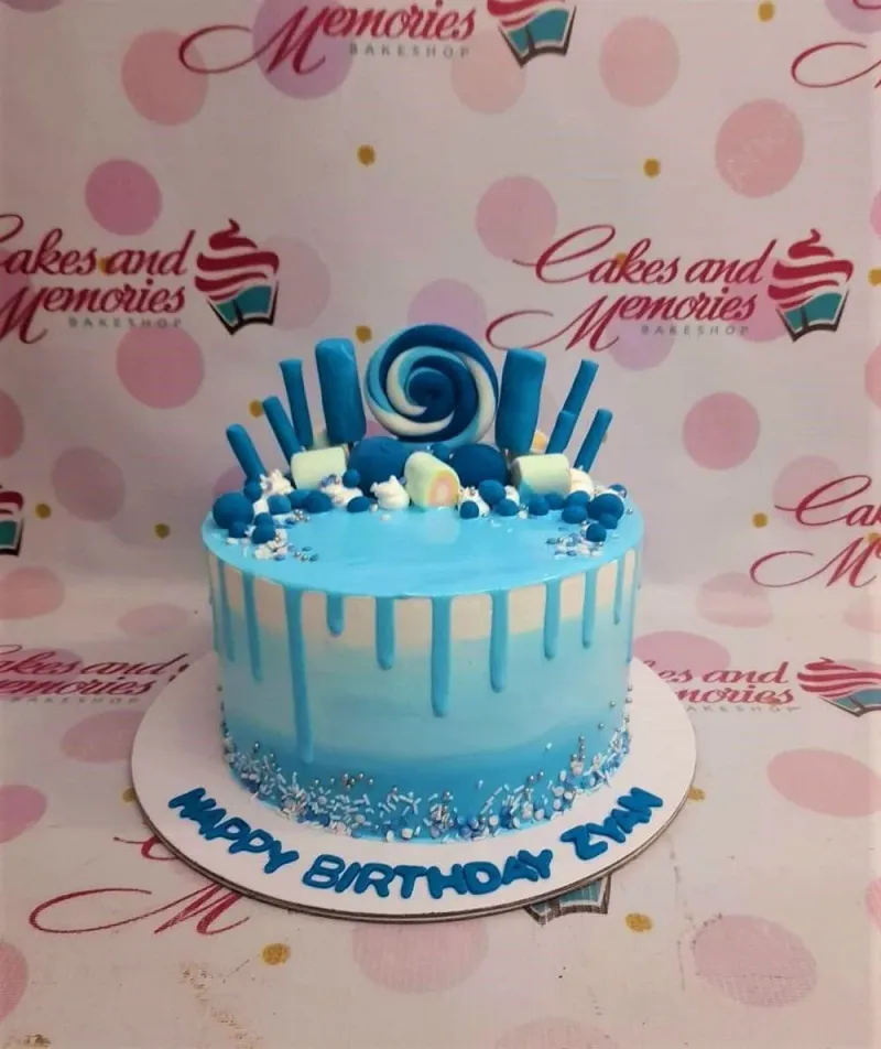 A blue and white 1-tier Blue Drip cake, featuring a blue swirl lollipop, white marshmallows, blue fondant accents, and sprinkles. A blue and white 1-tier Blue Drip cake, featuring a blue swirl lollipop, white marshmallows, blue fondant accents, and sprinkles.
