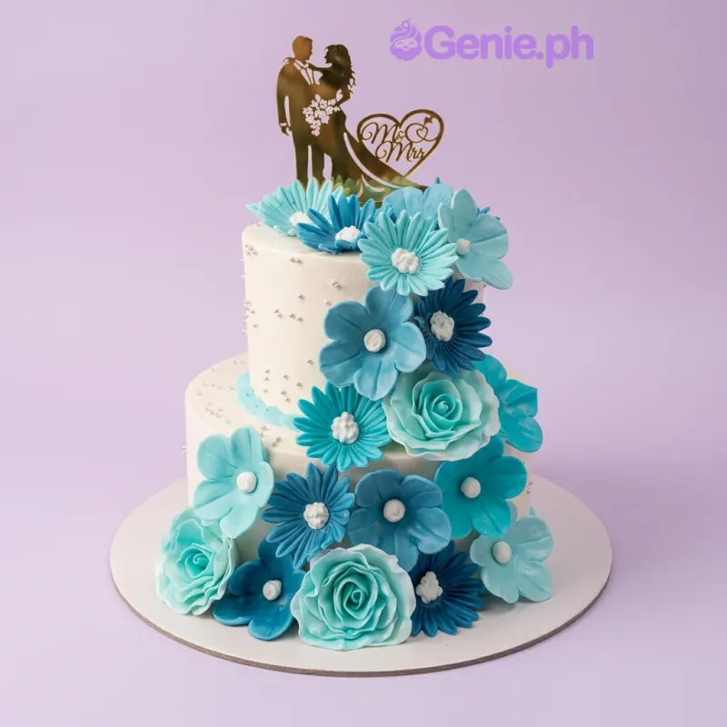 A white two-tier cake themed around Blue Floral Wedding, featuring blue and teal gumpaste flowers and a gold couple topper. A white two-tier cake themed around Blue Floral Wedding, featuring blue and teal gumpaste flowers and a gold couple topper.