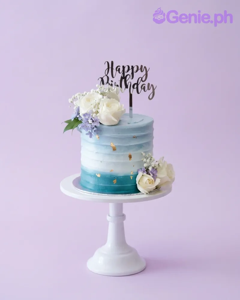 A blue and white ombre single-tier floral cake, featuring a 'Happy Birthday' script topper, white roses, lavender, and gold leaf. A blue and white ombre single-tier floral cake, featuring a 'Happy Birthday' script topper, white roses, lavender, and gold leaf.