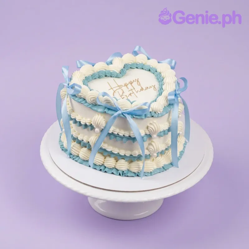 A white and light blue heart-shaped 1-tier cake themed 'Vintage Heart,' featuring ruffled icing borders, blue satin ribbons, and a 'Happy Birthday' message. A white and light blue heart-shaped 1-tier cake themed 'Vintage Heart,' featuring ruffled icing borders, blue satin ribbons, and a 'Happy Birthday' message.