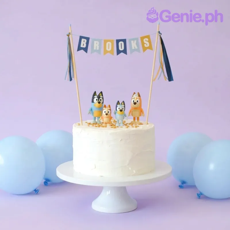 A white 1-tier cake themed around Bluey, featuring Bluey character toys, a 'Brooks' name banner, and golden chocolate drops. A white 1-tier cake themed around Bluey, featuring Bluey character toys, a 'Brooks' name banner, and golden chocolate drops.