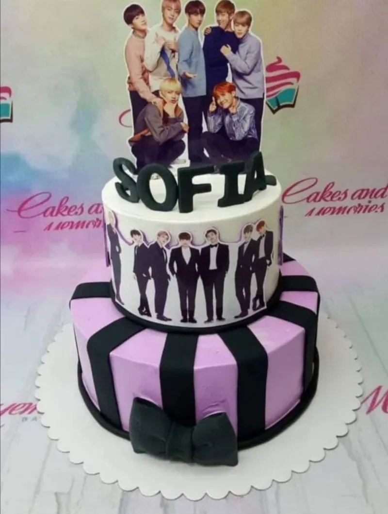 Two-tier purple and white BTS themed cake with black stripes, a large black bow, and printed character toppers. Two-tier purple and white BTS themed cake with black stripes, a large black bow, and printed character toppers.