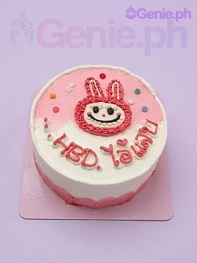 A pink and white 1-tier bunny-themed cake, featuring a hand-piped red bunny character, multicolored icing dots, and red Thai script. A pink and white 1-tier bunny-themed cake, featuring a hand-piped red bunny character, multicolored icing dots, and red Thai script.