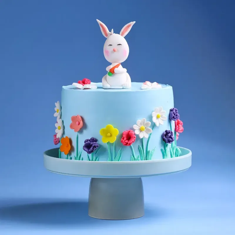 Light blue 1-tier cake with soft icing, a white bunny figurine holding a carrot, and colorful fondant flowers on the sides Light blue 1-tier cake with soft icing, a white bunny figurine holding a carrot, and colorful fondant flowers on the sides