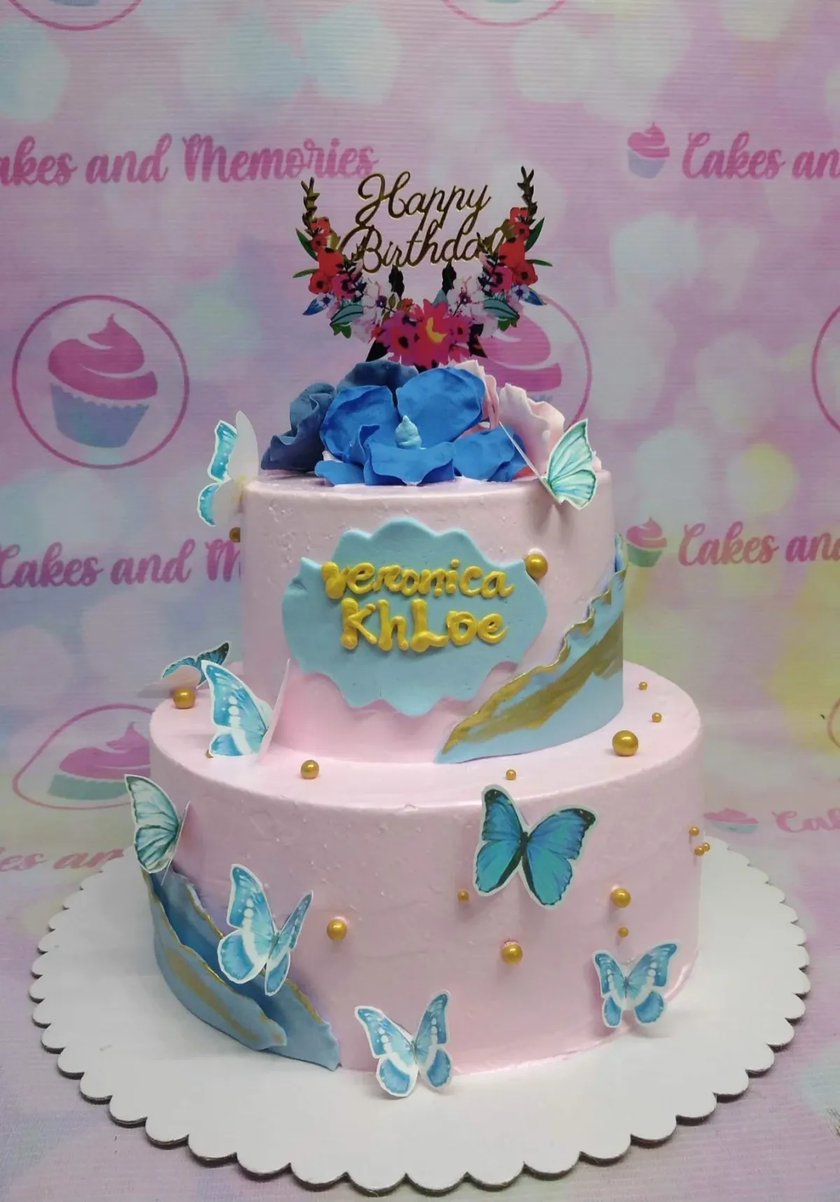Two-tier pink birthday cake decorated with blue butterflies, gold dragees, and a floral Happy Birthday topper. Two-tier pink birthday cake decorated with blue butterflies, gold dragees, and a floral Happy Birthday topper.