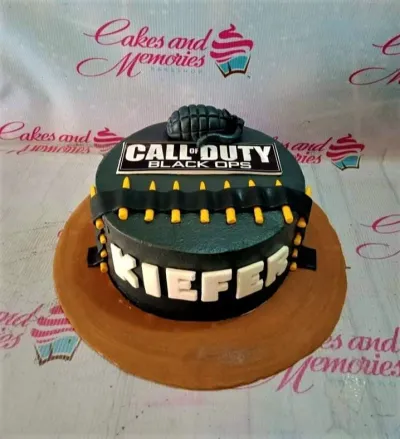 A black 1 Tier Fondant Call Of Duty Black Ops cake, featuring a grenade, game logo, bullet belt, and white 'KIEFER' text. A black 1 Tier Fondant Call Of Duty Black Ops cake, featuring a grenade, game logo, bullet belt, and white 'KIEFER' text.