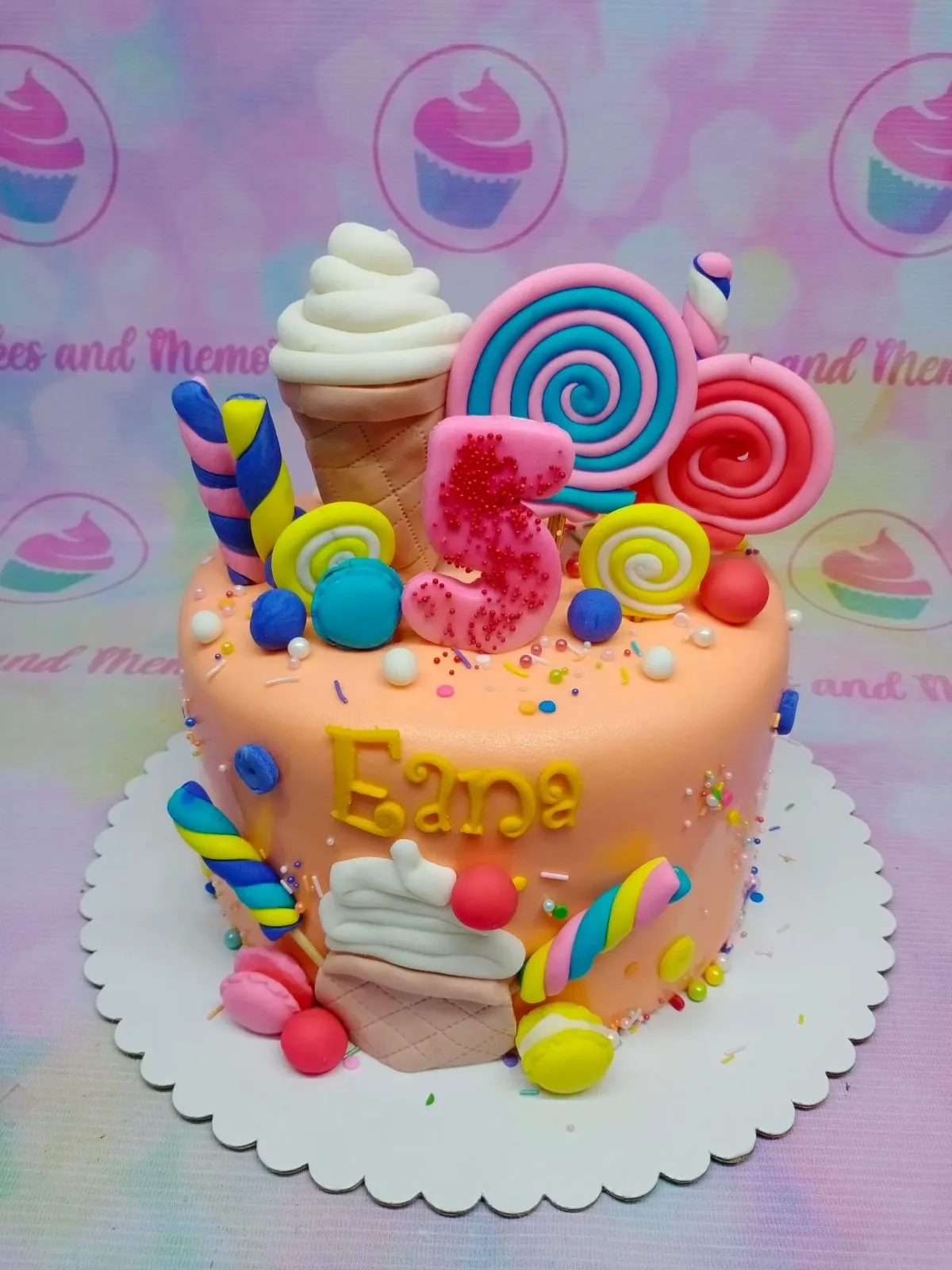 Peach-colored candy-themed birthday cake with fondant lollipops, ice cream cones, macarons, and a pink glittery number 5 topper. Peach-colored candy-themed birthday cake with fondant lollipops, ice cream cones, macarons, and a pink glittery number 5 topper.