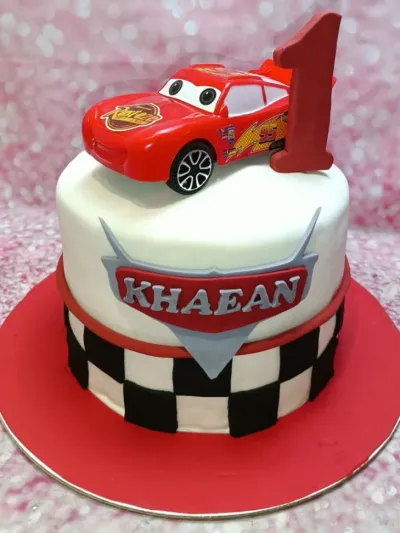 A white fondant Cars themed cake with a red Lightning McQueen toy car, a red number one, and a checkered racing flag pattern. A white fondant Cars themed cake with a red Lightning McQueen toy car, a red number one, and a checkered racing flag pattern.