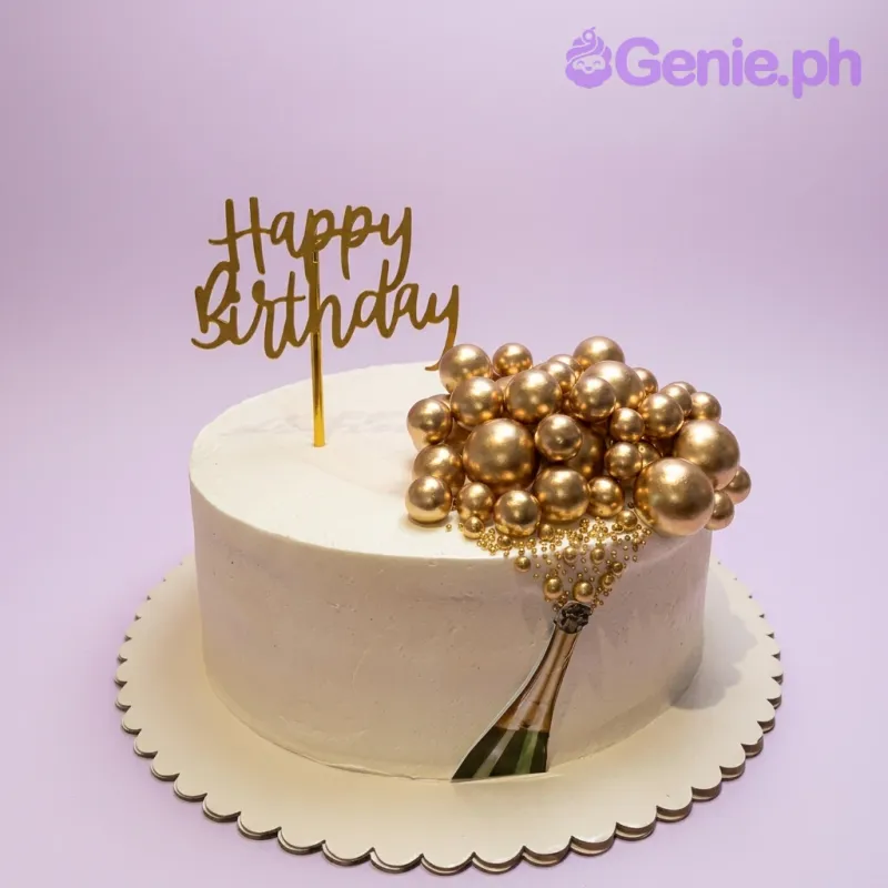 A white 1 Tier cake featuring a gold 'Happy Birthday' topper, gold decorative spheres, a champagne bottle printout, and gold sugar beads. A white 1 Tier cake featuring a gold 'Happy Birthday' topper, gold decorative spheres, a champagne bottle printout, and gold sugar beads.