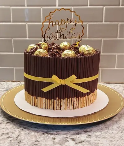 A round chocolate cake with chocolate biscuit sticks on the sides, topped with Ferrero Rocher chocolates and a gold Happy Birthday topper. A round chocolate cake with chocolate biscuit sticks on the sides, topped with Ferrero Rocher chocolates and a gold Happy Birthday topper.