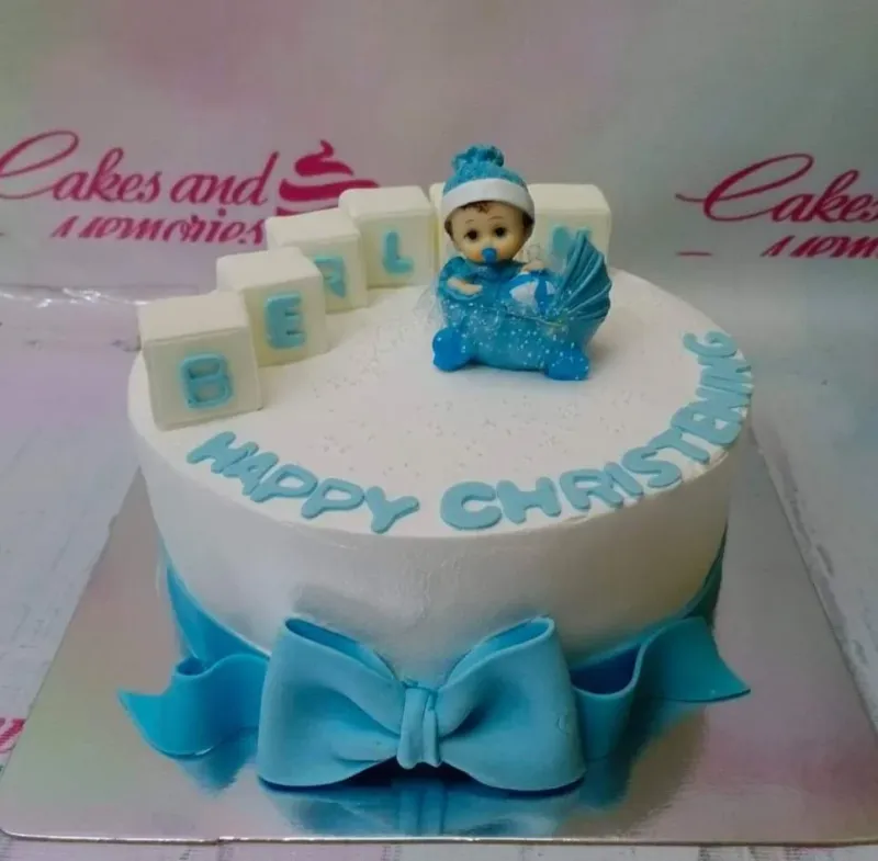 A white round 1-tier cake themed around christening, featuring a blue baby in a stroller topper, blue bow, and block letters spelling BERL. A white round 1-tier cake themed around christening, featuring a blue baby in a stroller topper, blue bow, and block letters spelling BERL.