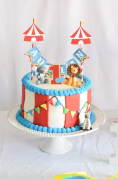White 1-tier circus theme cake with soft icing, red and white stripes, circus tent toppers, and animal figurines White 1-tier circus theme cake with soft icing, red and white stripes, circus tent toppers, and animal figurines