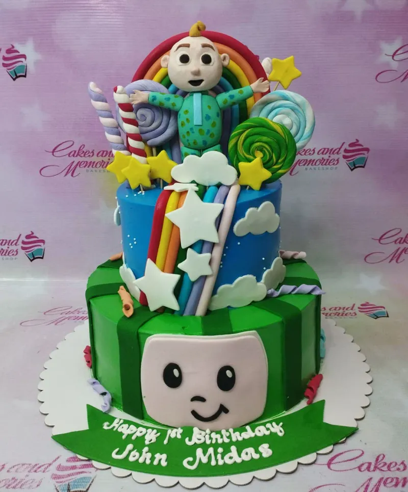 Two-tier Cocomelon themed birthday cake with a 3D JJ figure, rainbow, lollipops, and a green watermelon base tier. Two-tier Cocomelon themed birthday cake with a 3D JJ figure, rainbow, lollipops, and a green watermelon base tier.