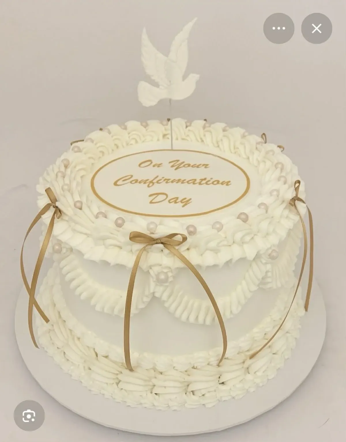 A white and gold 1-tier confirmation cake, featuring a white dove topper, gold ribbons, and ruffle piping. A white and gold 1-tier confirmation cake, featuring a white dove topper, gold ribbons, and ruffle piping.