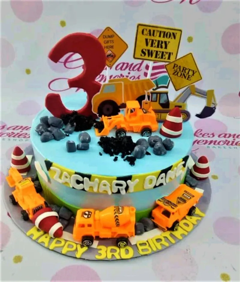 Light blue construction themed birthday cake with toy trucks, traffic cones, and a large red number three topper. Light blue construction themed birthday cake with toy trucks, traffic cones, and a large red number three topper.