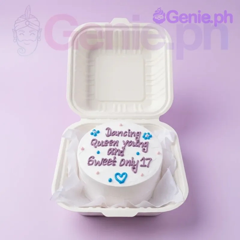 A white bento cake themed 'Dancing Queen', featuring a purple piped message, blue icing heart, and blue flowers. A white bento cake themed 'Dancing Queen', featuring a purple piped message, blue icing heart, and blue flowers.