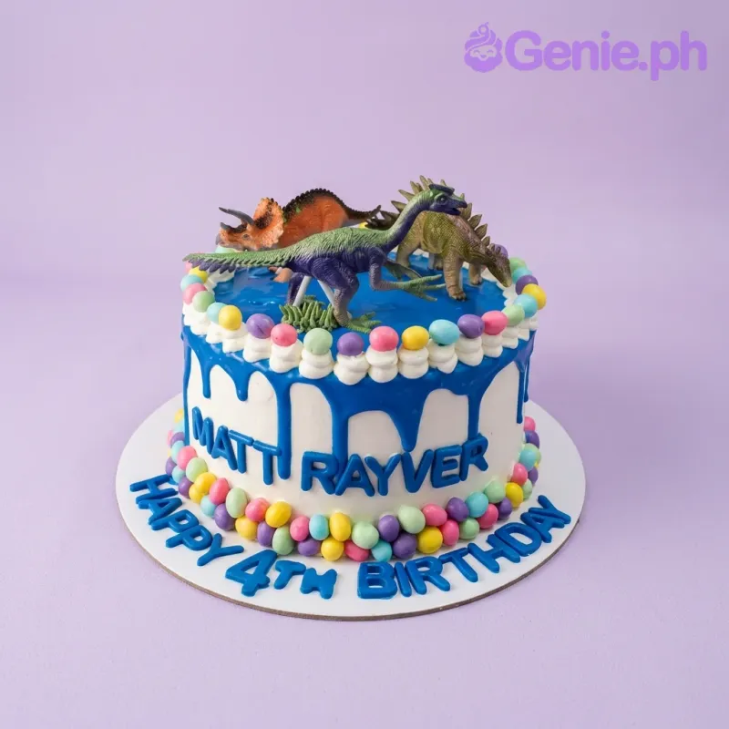 A white and blue 1 Tier cake themed around Dinosaurs, featuring three plastic dinosaur toy toppers and colorful candy borders. A white and blue 1 Tier cake themed around Dinosaurs, featuring three plastic dinosaur toy toppers and colorful candy borders.