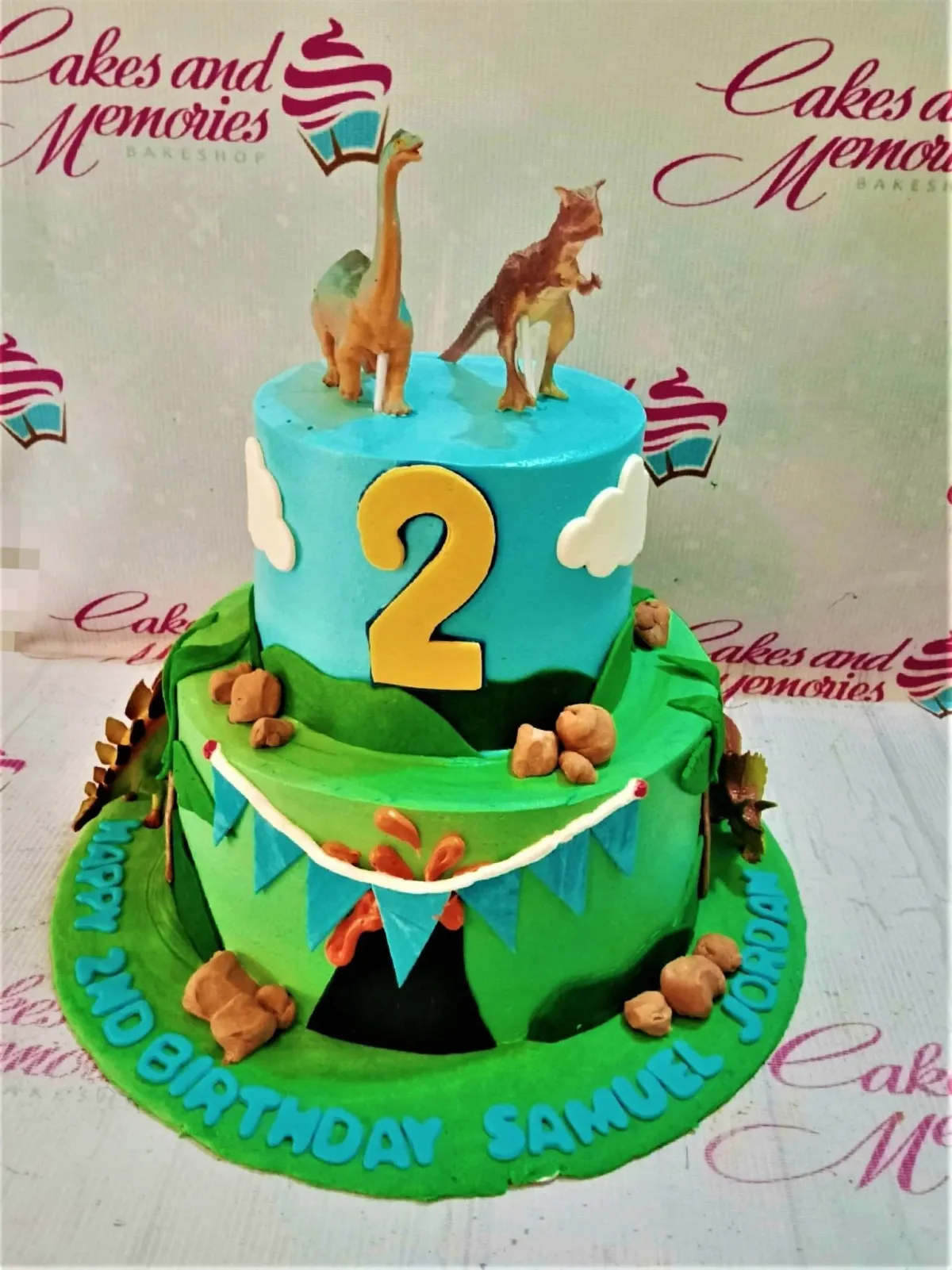 A blue and green two-tier cake themed around Dinosaurs, featuring 3D dinosaur figures, a volcano, and a yellow number 2. A blue and green two-tier cake themed around Dinosaurs, featuring 3D dinosaur figures, a volcano, and a yellow number 2.