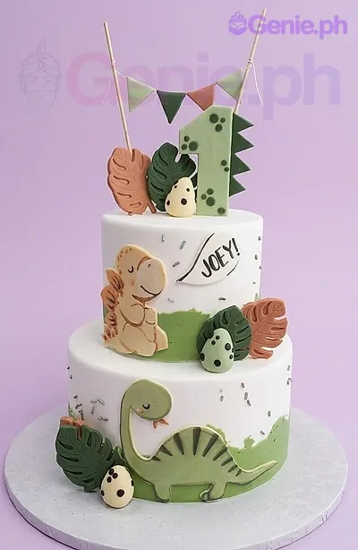 Two-tier white fondant dinosaur cake with green and brown gumpaste leaves, dinosaur eggs, and a large green number one topper. Two-tier white fondant dinosaur cake with green and brown gumpaste leaves, dinosaur eggs, and a large green number one topper.