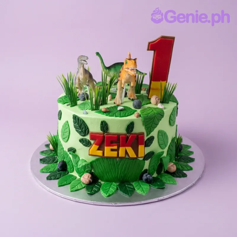 A green and red 1 Tier cake themed around Dinosaurs, featuring three dinosaur figures, a number one topper, fondant leaves, and 'ZEKI' in red letters. A green and red 1 Tier cake themed around Dinosaurs, featuring three dinosaur figures, a number one topper, fondant leaves, and 'ZEKI' in red letters.