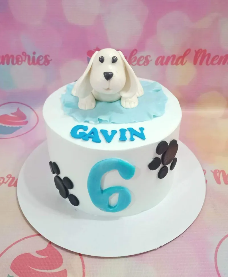A white round cake, dog themed, featuring fondant white dog, blue 'GAVIN' and '6', black paw prints. A white round cake, dog themed, featuring fondant white dog, blue 'GAVIN' and '6', black paw prints.
