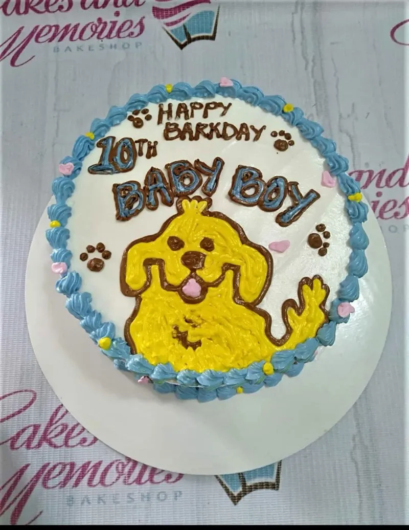 A white 1-tier round cake themed around Dog Birthday, featuring a yellow hand-piped dog character, blue rosettes, paw prints, hearts, and dots. A white 1-tier round cake themed around Dog Birthday, featuring a yellow hand-piped dog character, blue rosettes, paw prints, hearts, and dots.