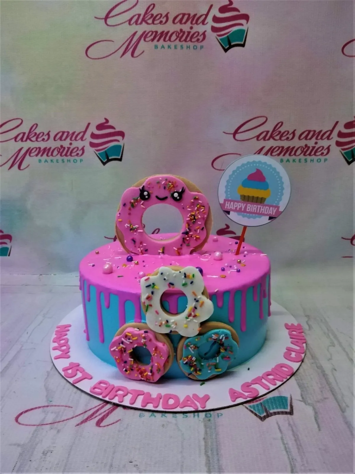 A pink and blue 1-tier Donut Birthday cake, featuring edible donut toppers, a pink drip, sprinkles, and 'HAPPY 1ST BIRTHDAY ASTRID CLAIRE' text. A pink and blue 1-tier Donut Birthday cake, featuring edible donut toppers, a pink drip, sprinkles, and 'HAPPY 1ST BIRTHDAY ASTRID CLAIRE' text.