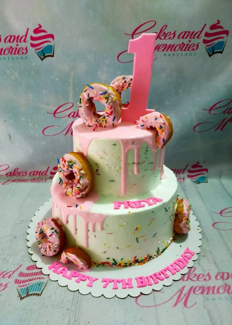 A white two-tier Donut cake with pink drip, colorful sprinkles, pink glazed donut toppers, and '7th Birthday' text. A white two-tier Donut cake with pink drip, colorful sprinkles, pink glazed donut toppers, and '7th Birthday' text.