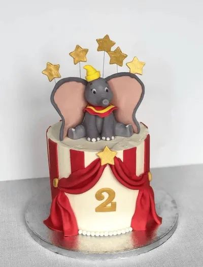 A red, white, and gold 1 Tier cake themed around Dumbo Circus, featuring a 3D Dumbo figure, gold stars, red and white stripes, and red fondant curtains. A red, white, and gold 1 Tier cake themed around Dumbo Circus, featuring a 3D Dumbo figure, gold stars, red and white stripes, and red fondant curtains.