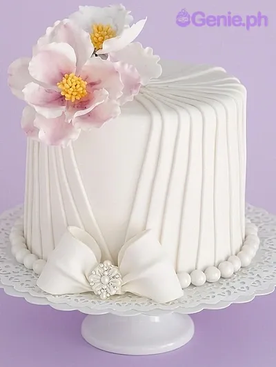 A white fondant cake themed Elegant Floral, featuring pink and white sugar peonies, pleated icing design, and a large white bow. A white fondant cake themed Elegant Floral, featuring pink and white sugar peonies, pleated icing design, and a large white bow.