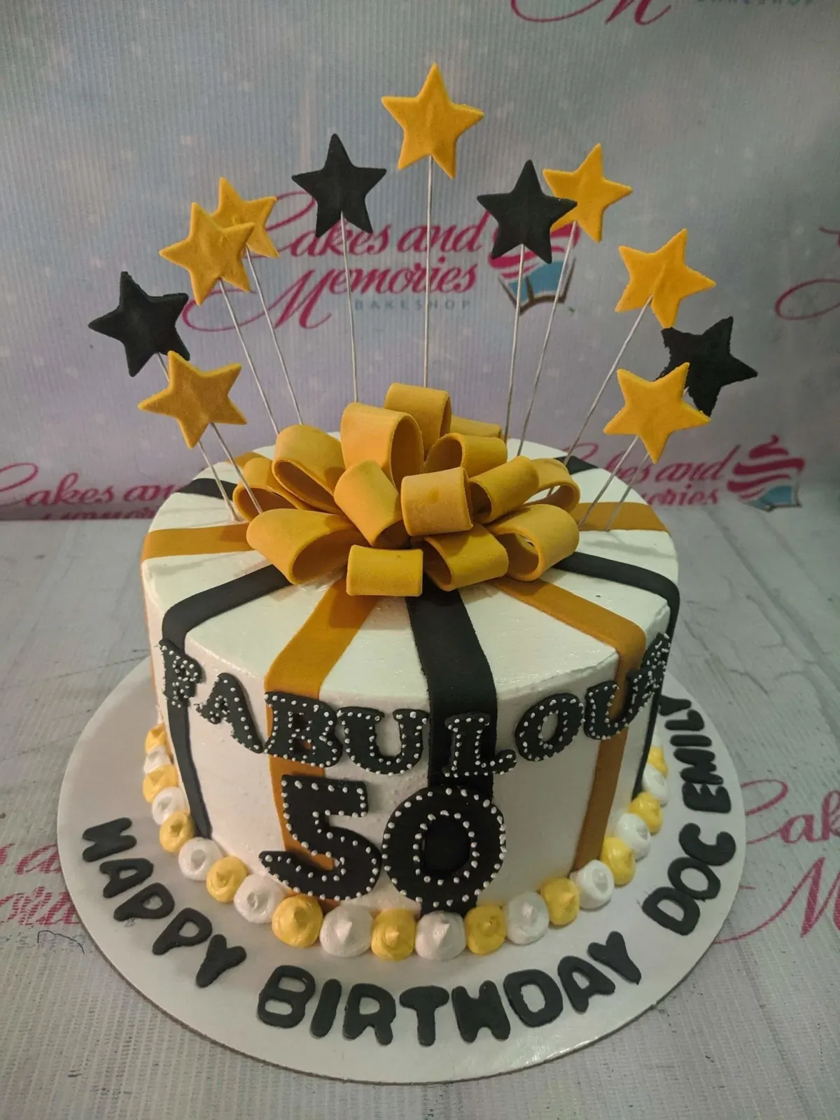 A white, black, gold 1-tier 'Fabulous 50' cake, featuring a gold bow, star toppers, and black and gold stripes. A white, black, gold 1-tier 'Fabulous 50' cake, featuring a gold bow, star toppers, and black and gold stripes.