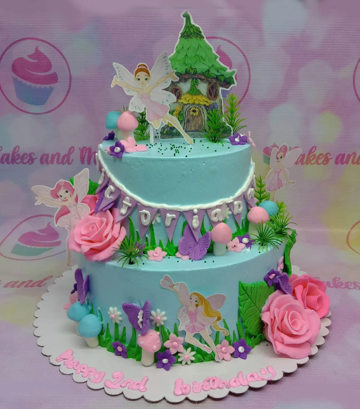 A light blue two-tier cake themed around Fairy Garden, featuring fairy house and character printouts, pink roses, and mushrooms. A light blue two-tier cake themed around Fairy Garden, featuring fairy house and character printouts, pink roses, and mushrooms.
