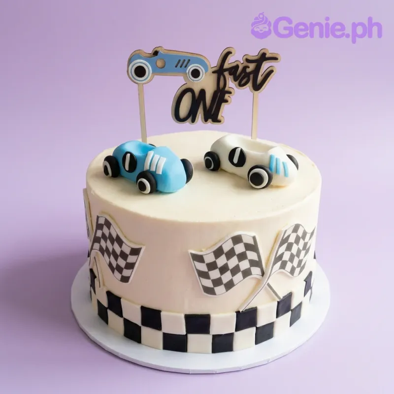 A light yellow single-tier cake with a Fast One Racing theme, featuring blue and white toy cars and checkered flag accents. A light yellow single-tier cake with a Fast One Racing theme, featuring blue and white toy cars and checkered flag accents.