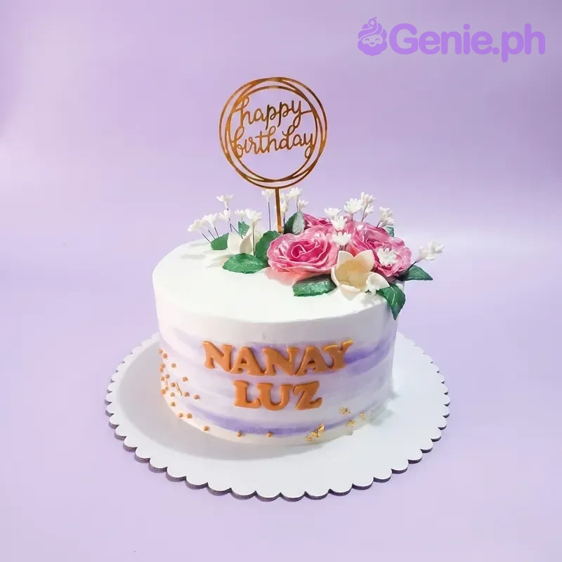 A white and purple round Floral Birthday cake, featuring a gold Happy Birthday topper, pink gumpaste roses, and gold lettering.