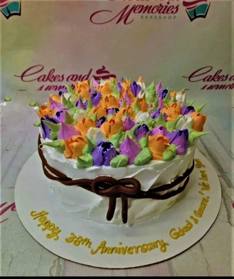 A white 1-tier Floral Anniversary cake, featuring purple, orange, and white icing flowers, green leaves, and a brown icing bow. A white 1-tier Floral Anniversary cake, featuring purple, orange, and white icing flowers, green leaves, and a brown icing bow.