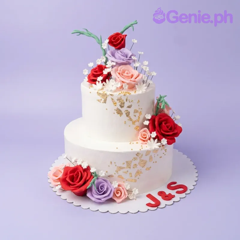 A white 2-tier cake themed Floral Anniversary, featuring red, purple, pink roses, gold leaf, and 'J&S' initials. A white 2-tier cake themed Floral Anniversary, featuring red, purple, pink roses, gold leaf, and 'J&S' initials.