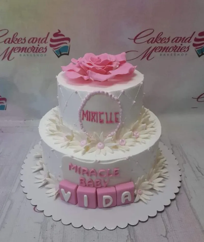 White 2-tier floral cake with pink fondant rose, daisy accents, and pink block letters for a baby celebration. White 2-tier floral cake with pink fondant rose, daisy accents, and pink block letters for a baby celebration.