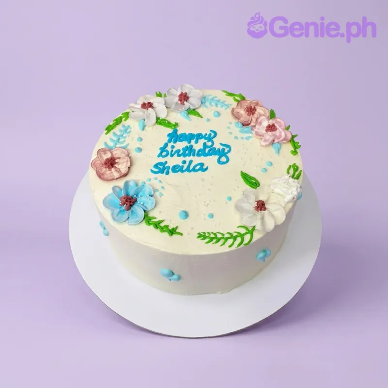 A white 1-tier cake themed around Floral Birthday, featuring colorful piped icing flowers and a blue 'Happy Birthday Sheila' message. A white 1-tier cake themed around Floral Birthday, featuring colorful piped icing flowers and a blue 'Happy Birthday Sheila' message.
