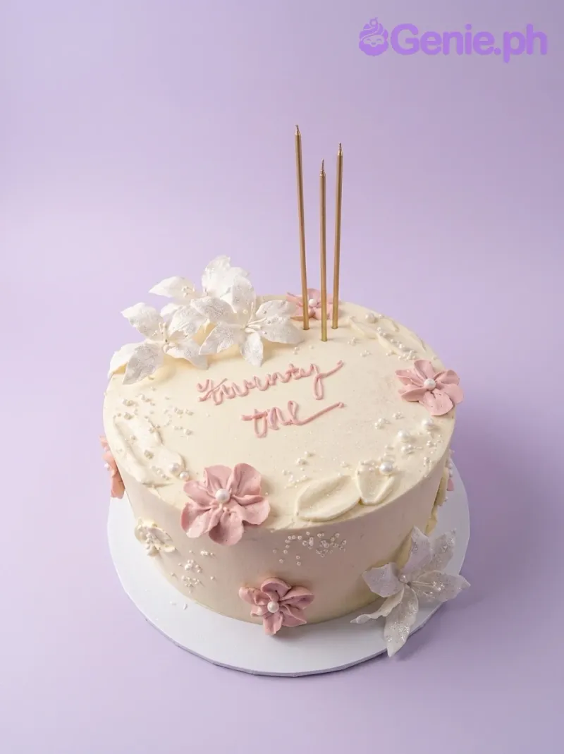 A white 1-tier cake themed Floral Birthday, featuring pink and white gumpaste flowers, gold candles, and pink icing text. A white 1-tier cake themed Floral Birthday, featuring pink and white gumpaste flowers, gold candles, and pink icing text.