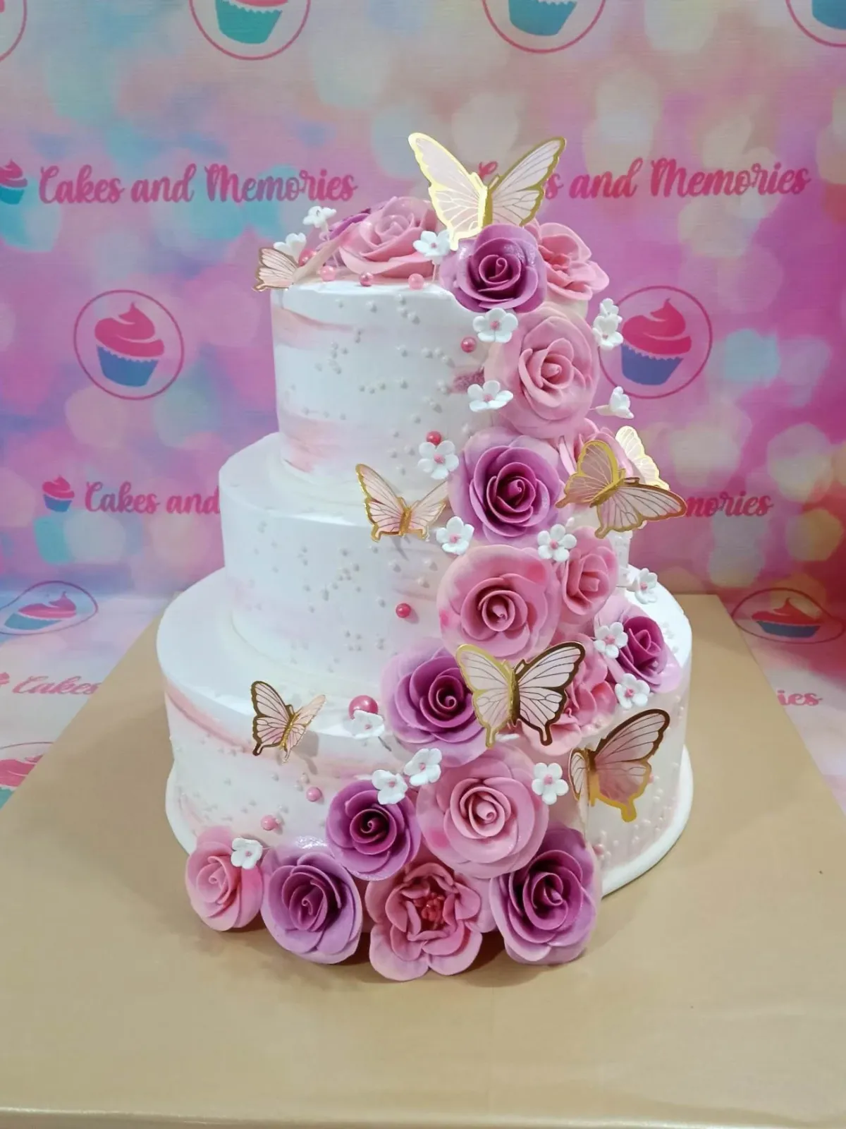 A white three-tier cake with a Floral Butterfly theme, featuring gold butterflies, pink and purple roses, and white rosettes. A white three-tier cake with a Floral Butterfly theme, featuring gold butterflies, pink and purple roses, and white rosettes.