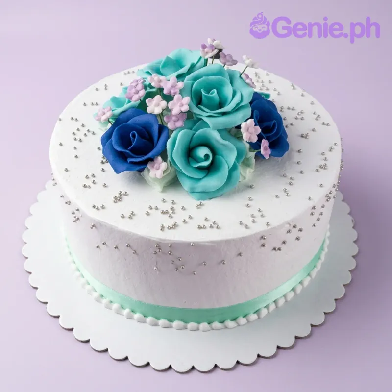A white 1-tier round cake themed Floral Celebration, with teal and navy blue gumpaste roses, purple filler flowers, and silver dragees. A white 1-tier round cake themed Floral Celebration, with teal and navy blue gumpaste roses, purple filler flowers, and silver dragees.