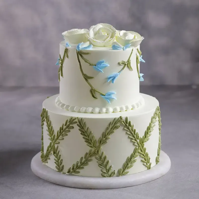 White 2-tier soft icing cake with piped green leaf lattice, blue floral vines, and white rose toppers White 2-tier soft icing cake with piped green leaf lattice, blue floral vines, and white rose toppers