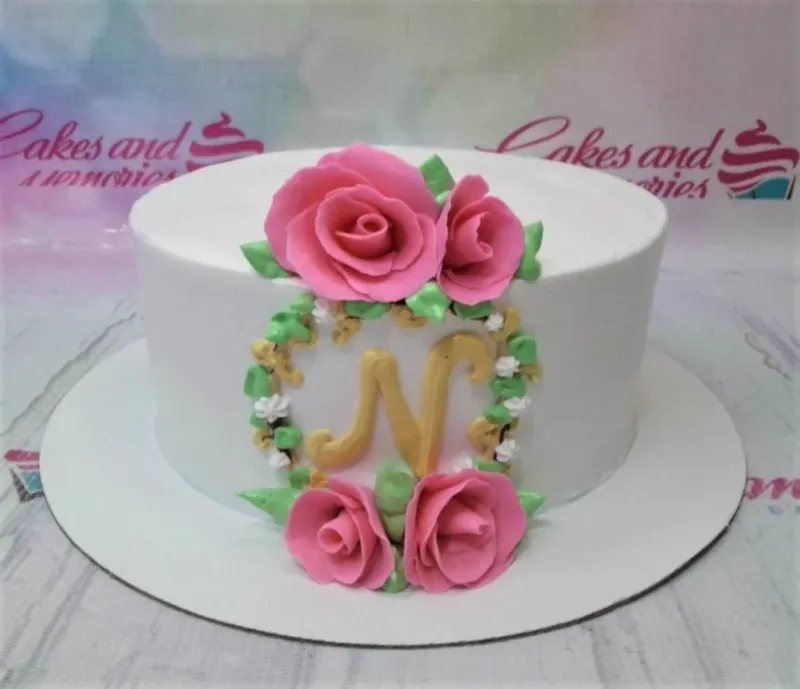 A white 1 Tier cake themed around Floral Monogram, featuring a gold letter N monogram, pink gumpaste roses, green leaves, and gold accents. A white 1 Tier cake themed around Floral Monogram, featuring a gold letter N monogram, pink gumpaste roses, green leaves, and gold accents.