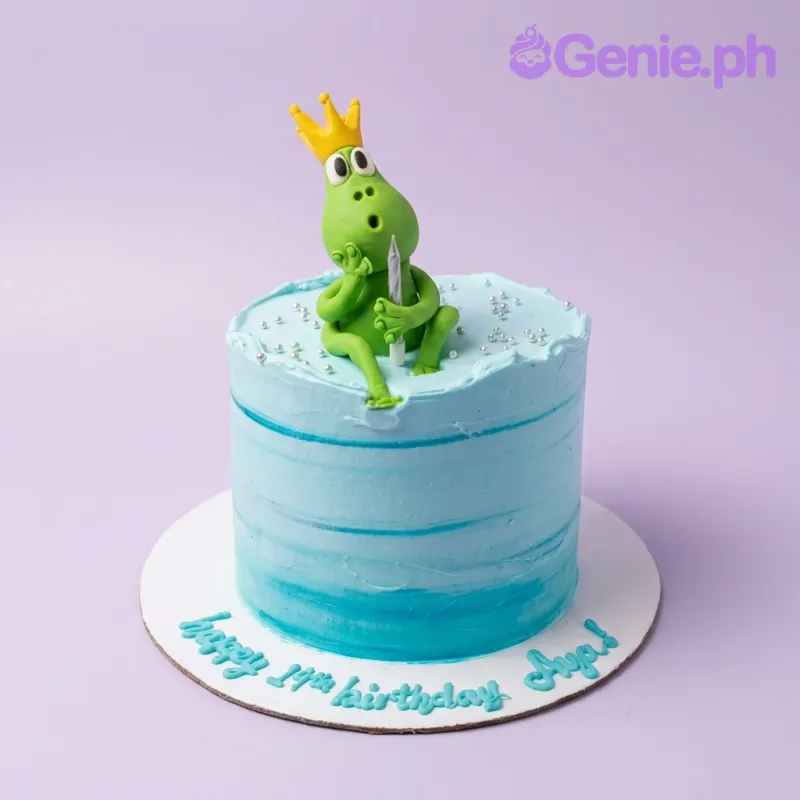 A light blue 1-tier Frog Prince cake with a green frog figurine wearing a yellow crown and silver dragees. A light blue 1-tier Frog Prince cake with a green frog figurine wearing a yellow crown and silver dragees.