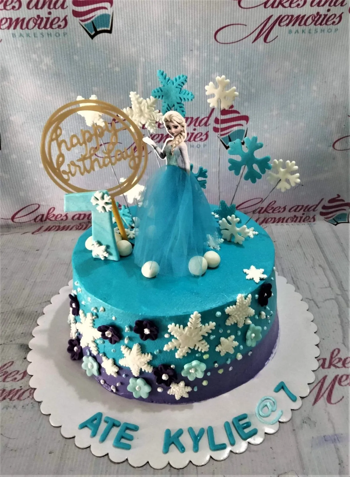 A blue and purple 1-tier cake themed around Frozen Elsa, featuring an Elsa printout, gold Happy Birthday sign, and number 7 topper, with snowflakes and purple flowers. A blue and purple 1-tier cake themed around Frozen Elsa, featuring an Elsa printout, gold Happy Birthday sign, and number 7 topper, with snowflakes and purple flowers.