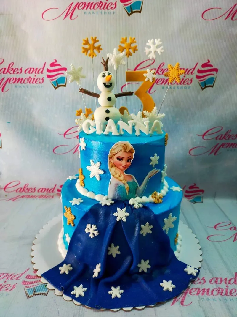 A blue two-tier cake themed Frozen Elsa, featuring Elsa and Olaf figures, a gold number three, dark blue drape, and white and gold snowflakes. A blue two-tier cake themed Frozen Elsa, featuring Elsa and Olaf figures, a gold number three, dark blue drape, and white and gold snowflakes.