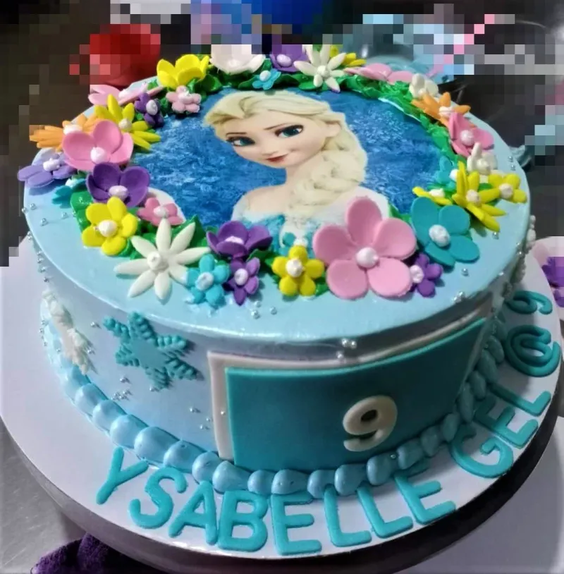 A light blue 1-tier cake themed around Frozen Elsa, featuring an Elsa printout and colorful fondant flowers with silver pearls. A light blue 1-tier cake themed around Frozen Elsa, featuring an Elsa printout and colorful fondant flowers with silver pearls.